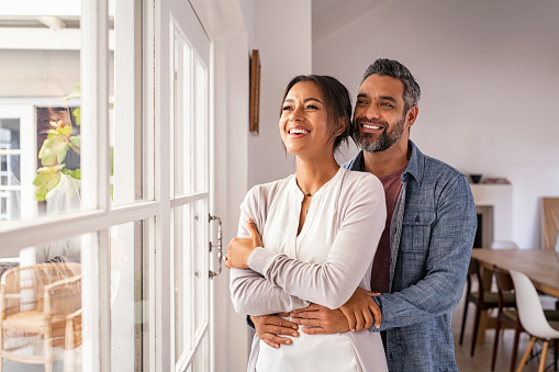 Smiling mid adult couple hugging each other and standing near window while looking outside. Happy and romantic mature man embracing hispanic wife from behind while standing at home with copy space. Future, vision and daydream concept.